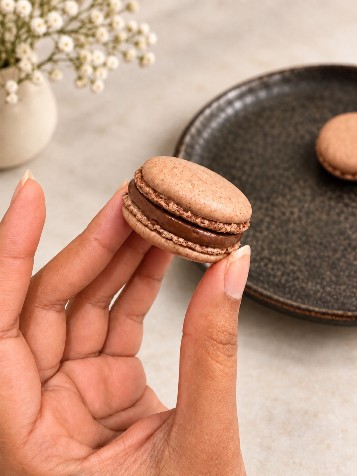 A chocolate macaron held between finger and thumb, its ganache centre showing